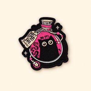 May include: An embroidered patch featuring a pink potion bottle with a black cat inside. The bottle has a cork stopper and a beige tag that reads DON'T OPEN EVIL inside. Above the cat, the text DO IT! is embroidered in black thread.