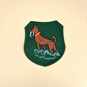 May include: Embroidered patch featuring a brown dog with a red collar, holding a blue object in its mouth, set against a green shield-shaped background. The dog stands above a stylized blue water design. The patch is likely for clothing or accessories.