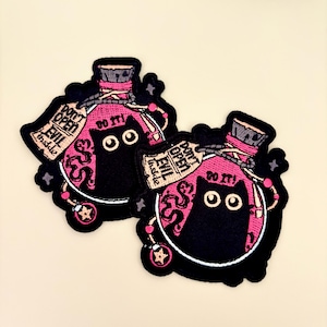 May include: Two embroidered patches featuring a black cat inside a pink potion bottle. The patches have the text "DON'T OPEN EVIL inside" and "DO IT!". The patches are outlined in black and have a light beige background.