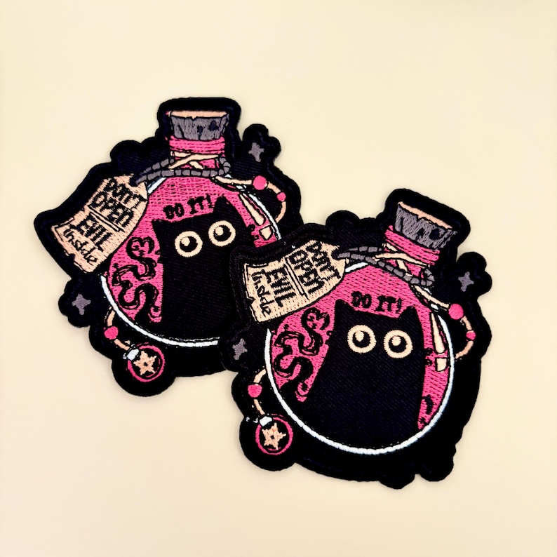 May include: Two embroidered patches featuring a black cat inside a pink potion bottle. The patches have the text "DON'T OPEN EVIL inside" and "DO IT!". The patches are outlined in black and have a light beige background.