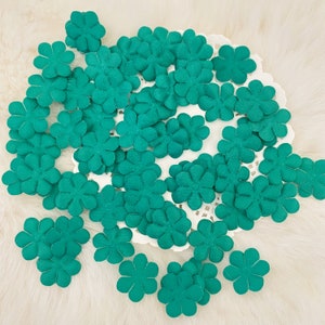 May include: A collection of small, teal-colored fabric flower embellishments. The flowers are arranged on a white, lacy doily, creating a delicate and craft-focused aesthetic. Ideal for scrapbooking, card making, or other DIY projects.