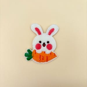 May include: Embroidered patch featuring a white bunny with pink inner ears and rosy cheeks, holding an orange carrot with green leaves. The patch is designed with a cute, cartoonish style.