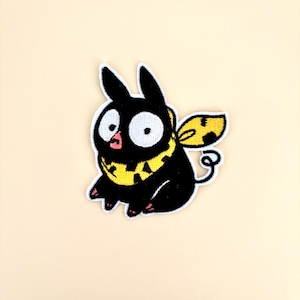 May include: An embroidered patch featuring a black creature with large white eyes, a small pink mouth, and a yellow scarf with black spots. The creature has pointed ears, a yellow wing with black spots, and a small curly tail on a white border.