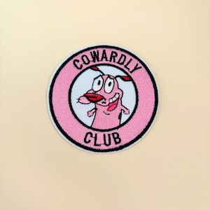 May include: A round, embroidered patch featuring the cartoon dog Courage from the show "Courage the Cowardly Dog." The patch has a pink border with the words "Cowardly Club" in black. Courage is smiling with his tongue out.