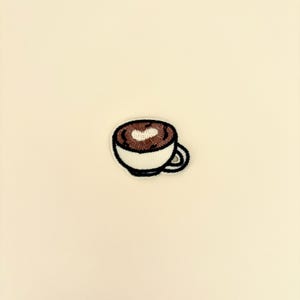 Peut inclure: Écusson brodé d'une tasse de café avec un motif en forme de cœur dans la mousse. La tasse est blanche avec un motif de café brun et un contour noir. L'écusson est sur un fond crème.