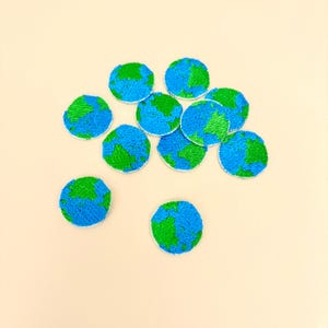 May include: A collection of embroidered patches shaped like the Earth. Each patch features a blue ocean and green landmass design. The patches are round with a white border and are scattered on a light beige surface.