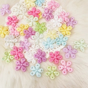 Könnte beinhalten: Sortiment an pastellfarbenen, gestickten Blumenverzierungen. Die Blumen sind in verschiedenen Schattierungen von Rosa, Blau, Gelb und Grün. Die Blumen sind auf einer weißen Spitzenunterlage angeordnet.