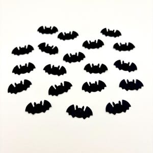 May include: A collection of nineteen small, flat, black bat-shaped cutouts arranged in a scattered pattern. Each individual piece features a rounded body with pointed ears and outstretched wings, creating a uniform silhouette for each bat shape.