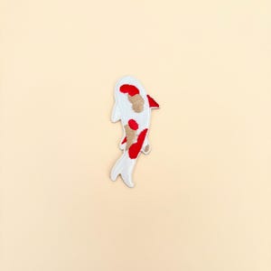 May include: Embroidered patch in the shape of a koi fish. The fish is white with red and tan markings. The patch is designed to be ironed on or sewn onto fabric. The koi fish is a symbol of good luck and perseverance.