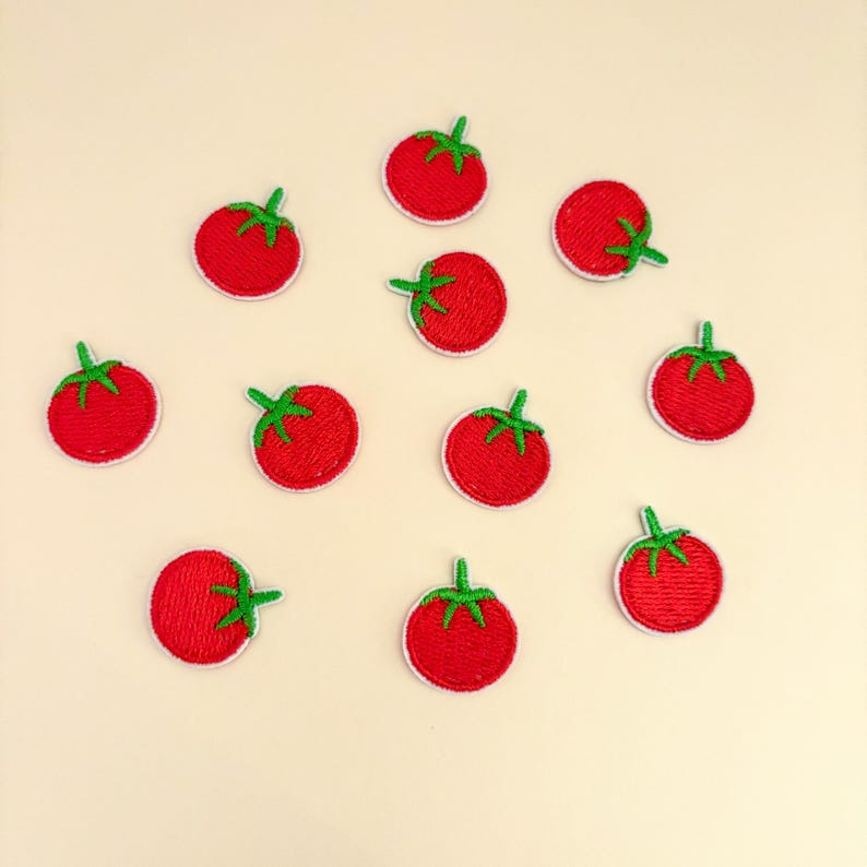 Pode incluir: Uma cole&ccedil;&atilde;o de onze patches bordados de tomate. Cada patch apresenta um tomate vermelho vibrante com um caule verde e uma borda branca. Os patches est&atilde;o espalhados em um fundo bege claro.