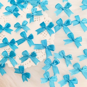 May include: A collection of small, light blue satin bows. The bows are arranged on a white doily and a white surface.