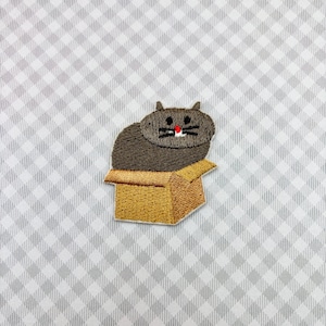 May include: Embroidered patch featuring a gray cat sitting in a cardboard box. The cat has black whiskers and a red nose. The box is tan with brown stitching. The background is a gray and white checkered pattern.