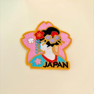 May include: Embroidered iron-on patch featuring a geisha in a pink cherry blossom design with the word "JAPAN" in yellow lettering.