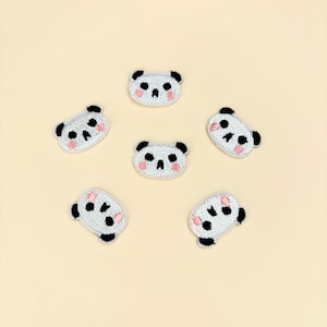 May include: Embroidered panda bear patches in a circular arrangement. Each patch features a white face with black eyes, ears, and nose, and pink cheeks. The patches are on a light beige background.
