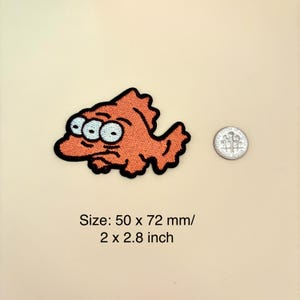 Op de afbeelding: Geborduurde oranje vispatch met drie ogen en een zwarte omtrek. De patch meet 50 x 72 mm. Een munt ligt naast de patch ter vergelijking van de grootte. De vis heeft een chagrijnige uitdrukking.