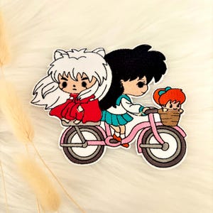 May include: Embroidered patch featuring three anime-style characters riding a pink bicycle. The characters have white, black, and orange hair, and are wearing red, blue, and white outfits. The bicycle has a brown basket.