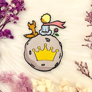 May include: Embroidered patch featuring a cartoon depiction of the Little Prince, a fox, and a rose on a planet with a gold crown. The patch has a white border and is set against a white, fluffy background.
