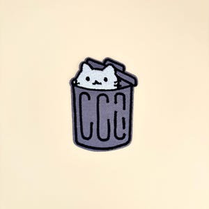 May include: Embroidered patch featuring a white cat peeking out of a gray trash can. The trash can has the letters "CCA" on the front. The patch is on a cream-colored background.
