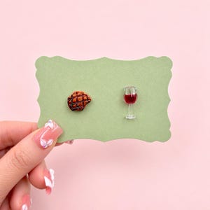 Peut inclure: Une paire de boucles d'oreilles clous représentant un steak grillé miniature avec des marques de cuisson foncées et un petit verre transparent rempli d'un liquide rouge foncé, fixés sur une carte de présentation vert sauge.