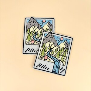 May include: Two embroidered patches in the shape of a Polaroid photo. Each patch features a mountain range, trees, a river, and a sun. The patches are in shades of blue, green, gray, and pink, with a white border.
