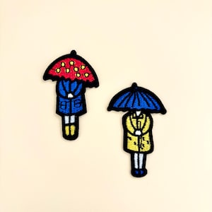 May include: Two embroidered patches featuring cartoon figures under umbrellas. One figure wears a blue coat and has a red umbrella with yellow dots. The other figure wears a yellow coat and has a blue umbrella.