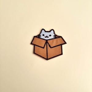 May include: Embroidered patch featuring a light blue cat peeking out of a brown cardboard box. The cat has a smiling face and black outlines. The box has a dark brown border and is set against a cream-colored background.