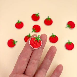 Pode incluir: Uma cole&ccedil;&atilde;o de pequenos patches de tomate bordados. Cada patch apresenta um tomate vermelho brilhante com uma haste verde e uma borda branca. Os patches est&atilde;o espalhados sobre um fundo bege claro.