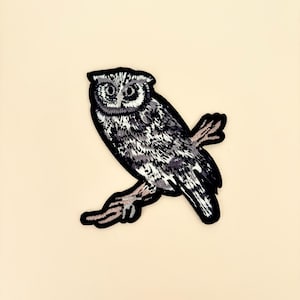 May include: An embroidered patch featuring a detailed owl perched on a branch. The owl is rendered in shades of gray and white, with a black outline. The patch is designed for application on clothing or accessories.