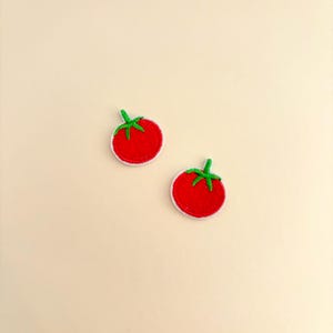 Pode incluir: Dois patches de tomate bordados. Cada patch apresenta um tomate vermelho brilhante com um caule verde e uma borda branca. Os patches s&atilde;o colocados em um fundo bege claro.