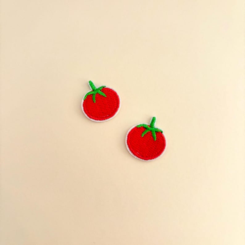 Pode incluir: Dois patches de tomate bordados. Cada patch apresenta um tomate vermelho brilhante com um caule verde e uma borda branca. Os patches s&atilde;o colocados em um fundo bege claro.