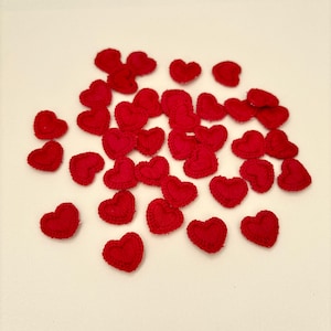 May include: A collection of small, red, heart-shaped fabric embellishments scattered on a white surface. The hearts are made of a textured material, possibly felt or a similar fabric, and are evenly distributed across the frame.