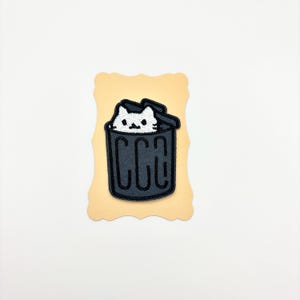 May include: An embroidered patch featuring a white cat with black facial details peeking out from inside a dark gray trash can with a lid. The design is rendered in a cartoon style with bold black outlines and is displayed on a light tan backing card.