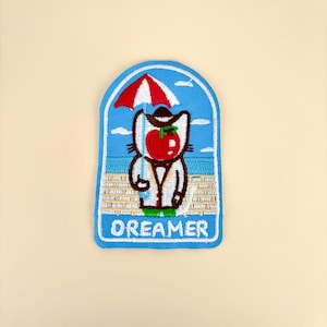 May include: An embroidered patch featuring a cat with a red apple head wearing a hat and coat, standing on a beach under a red and white striped umbrella. The background shows a blue sky with clouds and the word DREAMER in white capital letters.