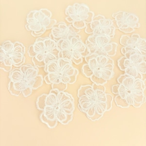 May include: A collection of white floral appliques featuring delicate embroidered outlines and sheer centers. Each piece is shaped like a five-petaled flower with intricate thread detailing, arranged in a scattered pattern across the frame.