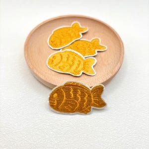 May include: Embroidered patches in the shape of a fish, in shades of yellow and gold, with white borders. Three patches are on a wooden plate, and one is in the foreground. The patches have detailed stitching.