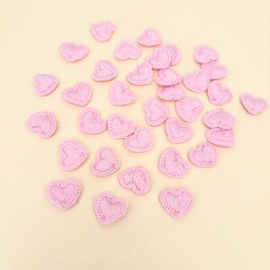 May include: A collection of small, pink, embroidered heart-shaped appliques scattered on a light yellow surface. Each heart has a detailed, stitched outline and a filled-in center, creating a textured appearance. These embellishments are ideal for crafts and DIY projects.