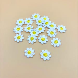 May include: A collection of small, embroidered daisy patches. Each patch features white petals surrounding a yellow center. The patches are scattered on a light orange background, suitable for crafting or embellishment projects.