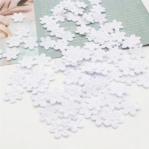 May include: A collection of small, white, daisy-shaped fabric embellishments scattered across a light-colored surface. The flowers have multiple petals and a textured appearance, suitable for crafts and decorations.