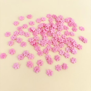 Peut inclure: Une collection de petites fleurs roses en tissu, dispersées sur une surface jaune clair. Les fleurs ont un aspect délicat et texturé, avec de multiples pétales et un centre légèrement surélevé. Ces fleurs décoratives sont adaptées aux travaux manuels.