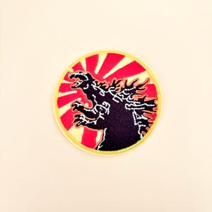 May include: A circular embroidered patch featuring a black silhouette of a monster with jagged dorsal fins. The background consists of alternating red and cream-colored rays radiating from the center, all enclosed by a yellow stitched border.