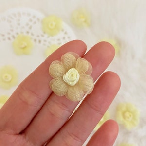 May include: A close-up of a pale yellow fabric flower with a smaller, lighter yellow fabric center. The flower is held in a hand against a white background.