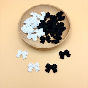 May include: A collection of white and black lace bows, some arranged in a wooden bowl, others scattered on a light orange surface. The bows are delicate and detailed, perfect for crafting or embellishing various items.