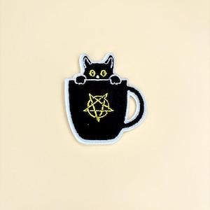May include: Embroidered patch featuring a black cat peeking over the rim of a black mug. The mug has a yellow pentagram design. The patch has a white border and is set against a cream-colored background.