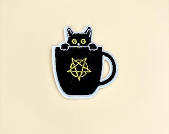 Puede incluir: Parche bordado con un gato negro asomándose por el borde de una taza negra. La taza tiene un diseño de pentagrama amarillo. El parche tiene un borde blanco y está sobre un fondo color crema.