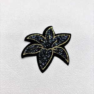 May include: Black flower-shaped applique with gold glitter accents. The applique has a black felt base and is covered in black sequins. The edges are outlined with gold glitter, creating a sparkling effect. This decorative applique can be used for various craft projects.