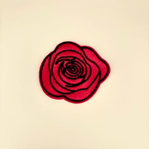 May include: A red embroidered rose patch with black outlines. The patch features a detailed design of a rose with multiple petals and a spiral center. The patch is likely for clothing or accessories.