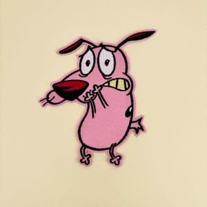 May include: An embroidered iron-on patch featuring a cartoon dog with pink fur, large white eyes, a dark red nose, and yellow teeth. The dog has a nervous expression with its paws held up to its face, black outlines, and a small black spot on its side.