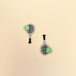 May include: Two embroidered patches featuring tennis rackets and tennis balls. The rackets are gray with black netting and black handles. The tennis balls are green. The patches are on a light beige background.