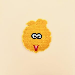 May include: A yellow embroidered patch featuring a stylized character face with a round, textured shape. The design includes large white eyes with black pupils, a small nose, and an open mouth showing a pink tongue, all outlined in yellow thread.