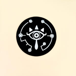 May include: A black circular patch featuring a white eye design with geometric shapes and lines. The eye has a central pupil, a triangular brow, and a teardrop shape below. The patch is on a cream-colored background.